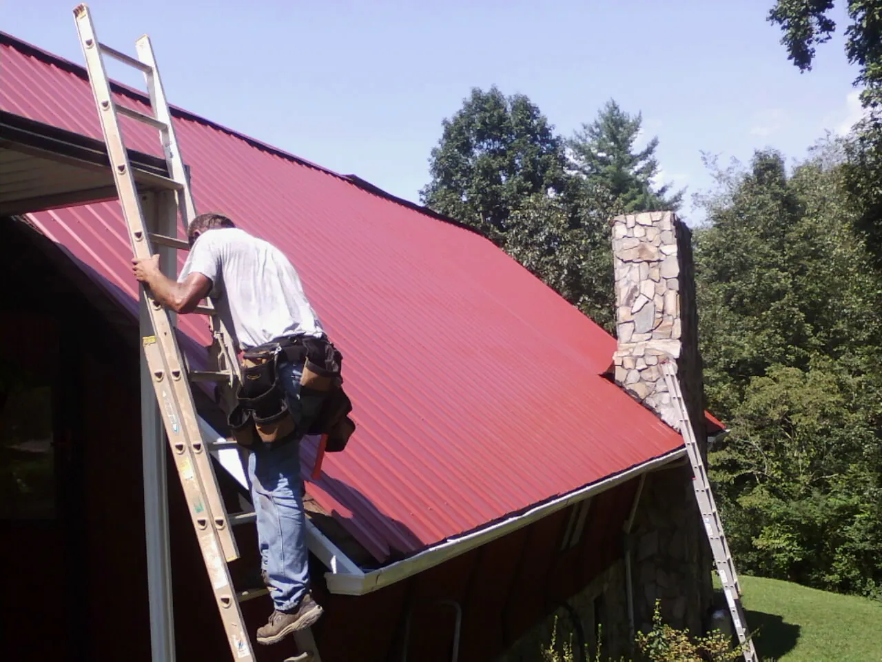 Roofer on a ladder beside a red metal roof and stone chimney — Soffit & Fascia Repair by Roofing Brothers in Kremmling